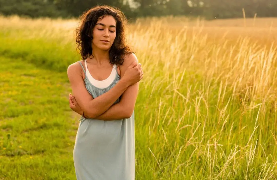 Woman stands confidently in a field, hugging herself gently with closed eyes. Hypnotherapy near Geebung. What Causes Painful Periods. Pyrrole Disorder. Brisbane Livewell Clinic
