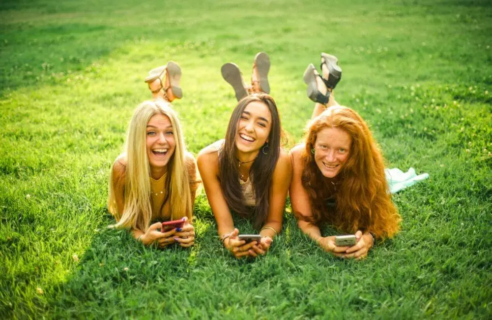 Three female friends lying on the grass while holding their smartphones. Hypnotherapy near Geebung. Hypnotherapy to Reduce Anxiety. Brisbane Livewell Clinic